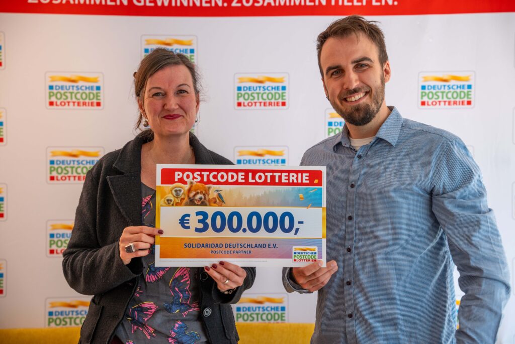 Zwei Vertreter:innen von Solidaridad Deutschland halten einen symbolischen Scheck über 300.000 Euro von der Deutschen Postcode Lotterie.