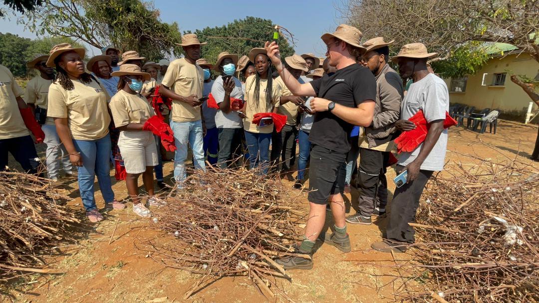 Eine Gruppe von Menschen in Outdoor-Arbeitskleidung und Hüten nimmt an einer landwirtschaftlichen Schulung im Freien teil, während ein Ausbilder zeigt, wie Pflanzenreste oder Äste für nachhaltige Bodenverbesserung vorbereitet werden.