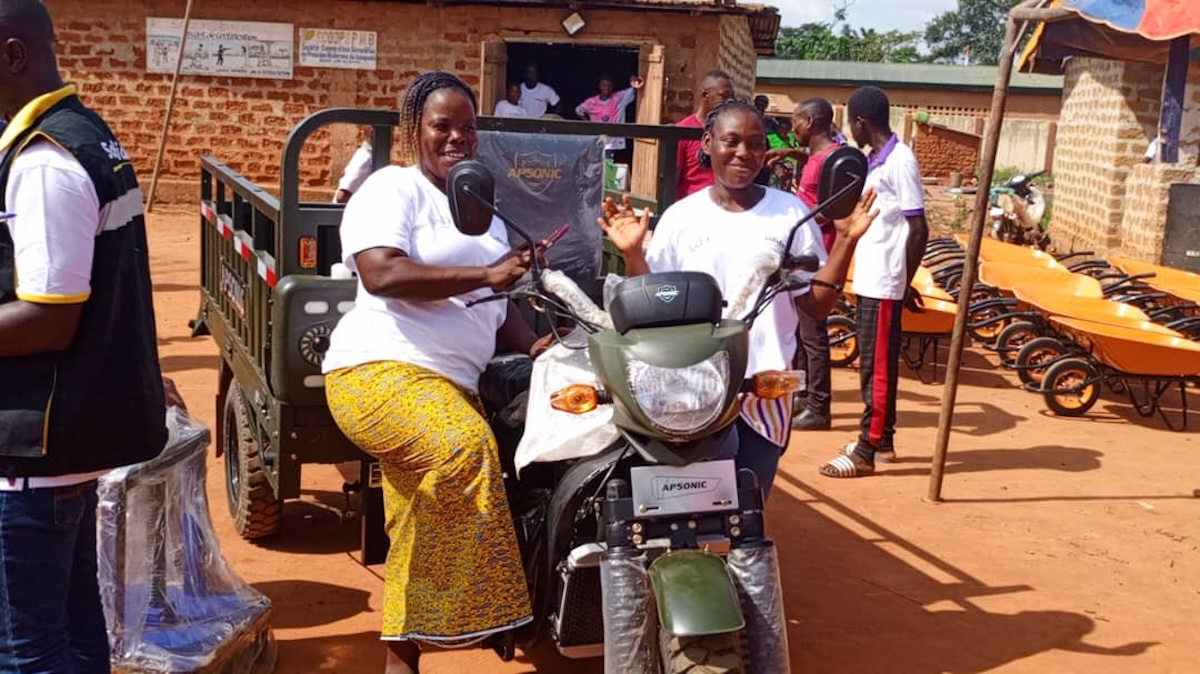 Auf dem Bild sind zwei Frauen mit einem Tricycle zu sehen. Sie erhalten dies im Rahmen des Projekts “CLIMATE-SMART COCOA”
