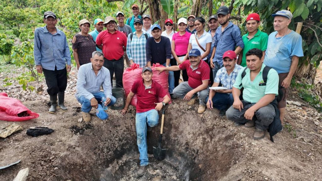 Eine Gruppe von Kakaobäuerinnen und Kakaobauern steht um eine Erdgrube mit handwerklich hergestellter Pflanzenkohle (Biochar) in einer Agroforst-Kakaoplantage in Nicaragua beim Pilotprojekt von Ritter Sport und Solidaridad.